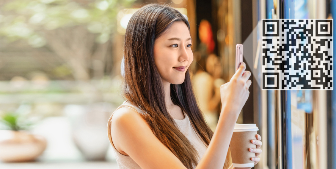 mulher jovem asiática sorrindo, escaneia um código QR com o celular na mão direita, enquanto segura café na outra